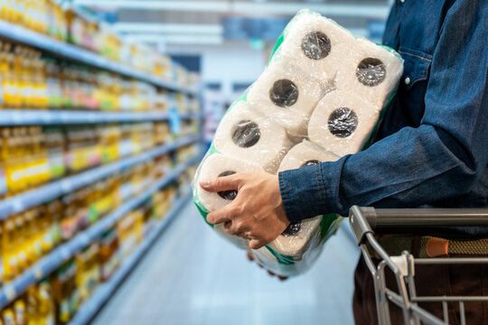 Shopper holding bulk packs of toilet paper beside a cart in a supermarket aisle, showing household supply shopping and preparation.