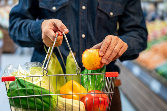 Close-up of a shopper in a dark jacket placing a red apple into a basket filled with fresh fruit and vegetables at a grocery store.