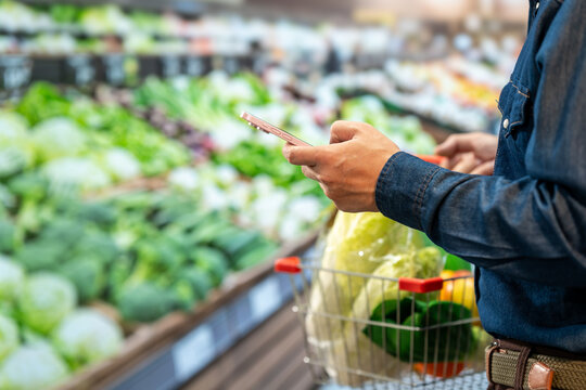 Shopper using a smartphone while choosing fresh vegetables in a supermarket produce aisle, with a basket of leafy greens and peppers.