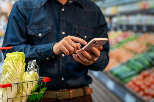 Man using a smartphone while shopping for fresh vegetables in a supermarket, with a cart holding cabbage and green onions.