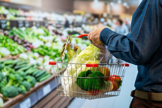 Close view of a shopper holding a basket filled with fresh vegetables while choosing produce in a bright supermarket grocery aisle.