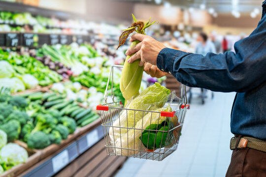 Shopper holding fresh corn over a basket filled with cabbage and green vegetables in a bright supermarket produce section.