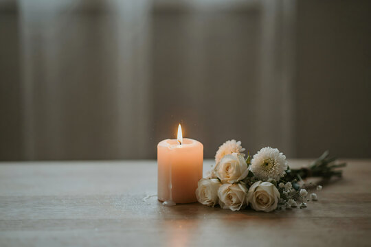 A single lit candle burning next to a small bouquet of white roses and chrysanthemums on a wooden table, soft lighting, peaceful atmosphere