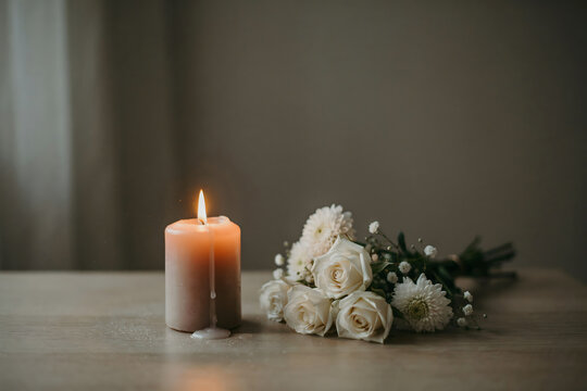 A single lit candle burning next to a small bouquet of white roses and chrysanthemums on a wooden table, soft lighting, peaceful atmosphere