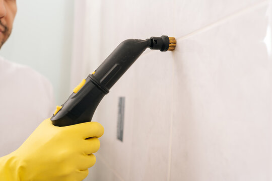 Male hand in yellow glove cleaning bathroom tiles and grout with powerful steam cleaner nozzle, ensuring deep hygiene and sanitation in domestic space. Concept of household chores and housework.