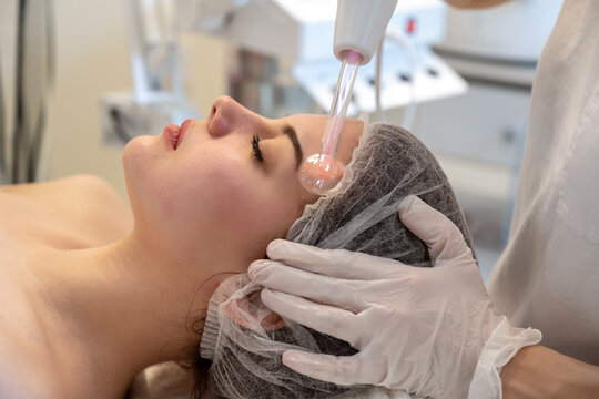Cosmetologist treating woman cheek skin with high frequency darsonval device