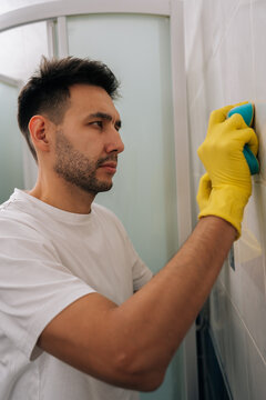Vertical portrait of man cleaning bathroom tiles with blue sponge while wearing yellow rubber gloves, focusing on scrubbing white tiled wall for maintaining hygiene and completing household chores.