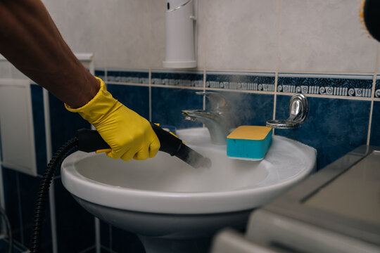 Person in yellow protective gloves deep cleaning bathroom sink using steam cleaner, generating visible steam while disinfecting surfaces and ensuring pristine hygiene in home, close-up.