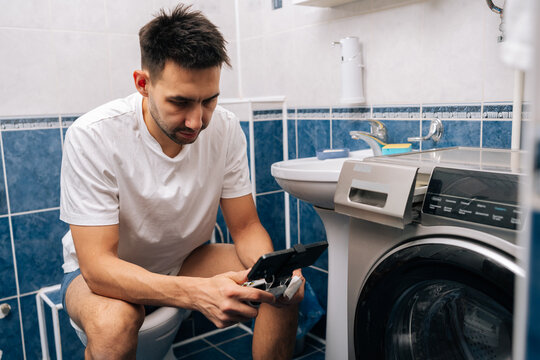 Handsome bearded young adult man in white t-shirt focusing on mobile gaming using smartphone and gamepad while sitting on toilet, portraying modern habits and technology integration in daily life.