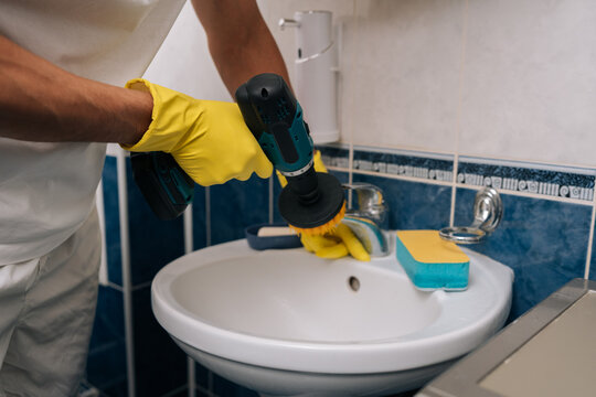 Cropped shot of man wearing yellow gloves using power drill fitted with brush attachment to quickly scrub and sanitize bathroom sink and faucet for efficient home cleaning and maintenance, close-up.