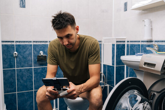 Man focusing on mobile gaming using smartphone and gamepad while sitting on toilet, portraying modern habits and technology integration in daily life.