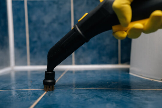Person in yellow protective gloves using powerful steam cleaner with brush attachment to deep clean blue ceramic bathroom tiles and grout lines, ensuring hygiene and sanitation, close-up.