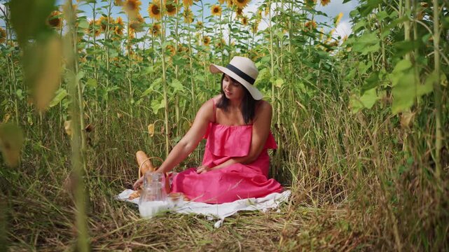 Woman in pink dress sits among sunflowers reaching for glass pitcher on white cloth with bread and jug nearby. Sunflower field surrounds her tall green stems fill frame creating vibrant