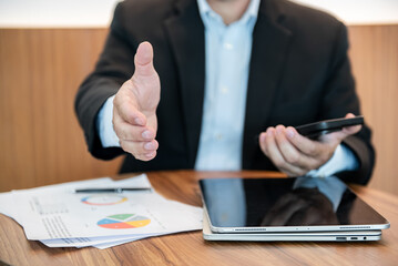 Businessman Extending Hand for Handshake at Office Desk with Tablet, Smartphone, and Financial Charts © BTM_Graphic