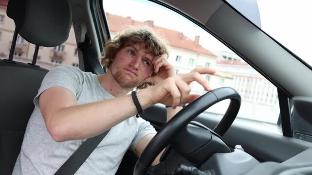 Bored male driver waits inside his parked car passing time with a tired expression while sitting behind the steering wheel and wondering when he can continue his day soon