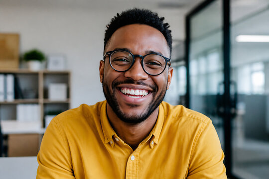 A smiling young Black man wearing glasses and a yellow shirt is pictured in a modern office setting.