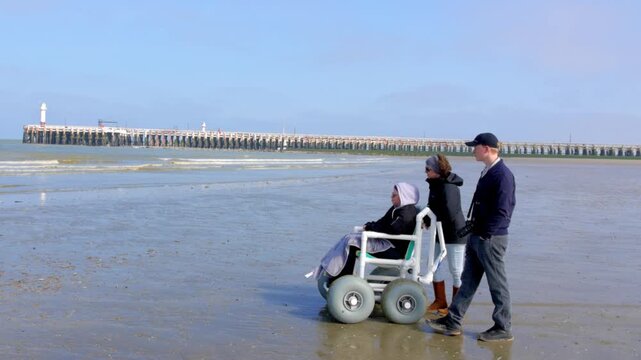 August 26, 2026 - Supportive Friends and Family Walking with a Person in an Accessible All-Terrain Wheelchair on a Sunny Beach