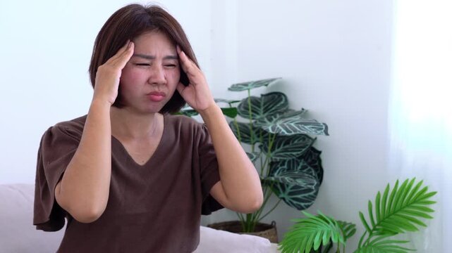 Asian woman holding her temples feeling severe headache, migraine pain, dizziness, or chronic stress while sitting indoors. Stressful life, healthcare, illness, and medical 