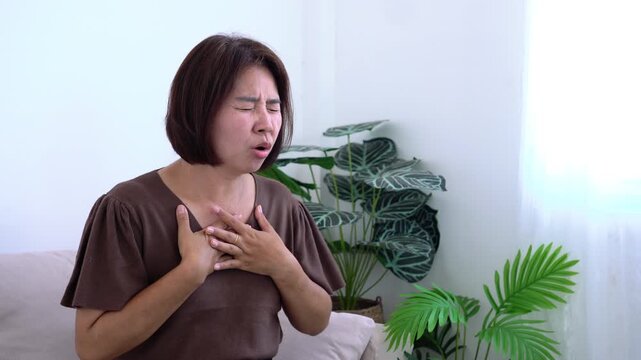 Middle-aged Asian woman holding her chest in pain, suffering from sudden chest tightness, panic attack, difficulty breathing, or heart attack symptom