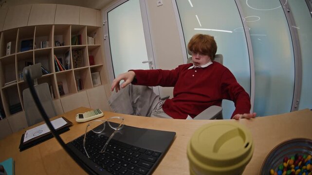 Caucasian young man with ginger hair typing on laptop, leaning back in office chair with feet up, relaxing and putting on glasses, fisheye lens. Footage suitable for youth entrepreneurship campaigns