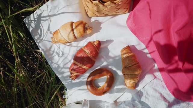 Close up of various baked goods arranged on white cloth outdoors. scene includes croissants bagels and bread in natural sunlight with shadows cast by surrounding grasses and objects. pink