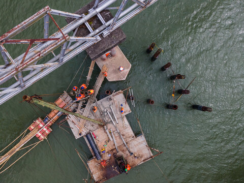 Aerial View of Construction Workers Wearing Orange Safety Jackets Installing Steel Stakes Across a River Beside Large Water Pipeline Infrastructure in Indonesia, Captured from Above.