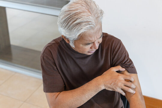 Southeast asian senior man scratching his itchy arm from a mosquito bite or skin rash at home, allergic reaction concept indoor scene