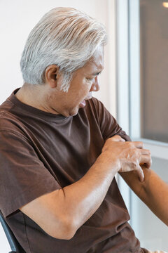 Southeast asian senior man scratching his itchy arm from a mosquito bite or skin rash at home, allergic reaction concept indoor scene