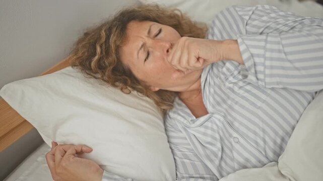 Woman lying on bed coughing into her hand with pillow visible in bedroom building; illness fatigue.
