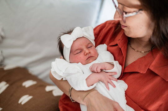 Mother holds her sleeping newborn baby dressed in a white outfit and headband with a large bow