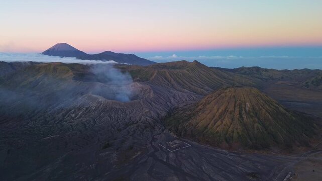 Drone pull-away over Mount Bromo during morning twilight, revealing the volcanic landscape, Mount Semeru and layered blue-to-pink sky above the Tengger caldera, Indonesia