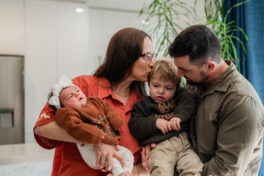 Mum kissing toddler sons head held by dad while carrying newborn baby girl