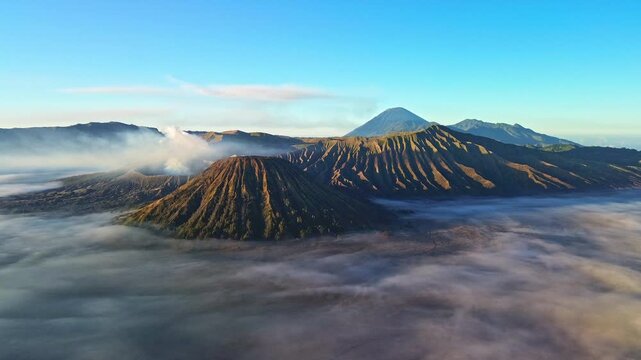 Slow elevated drone approach toward Mount Bromo from a distant, revealing an expansive sea of clouds, Mount Semeru and volcanic smoke beneath a blue sky, Indonesia