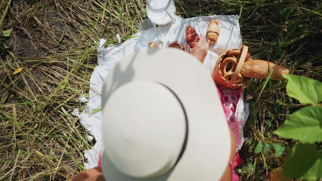 Woman in white hat enjoys selecting pastries from outdoor picnic spread on grass including croissants and baguettes. woven basket holds bread rolls next to blanket with glasses and milk jug