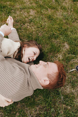Naklejka premium Smiling couple lying on grass, outdoor daylight, and cozy sweaters convey warmth, affection, and relaxed leisure, creating a candid lifestyle moment filled with happiness and connection on a natural,