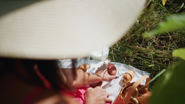Close up of woman in pink clothing reaching for pastries on white cloth spread outdoors. Various baked goods like croissants and bagels are arranged with sunlight casting shadows