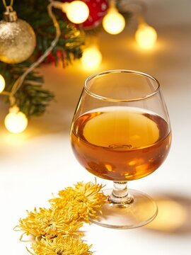 An elegant snifter glass of rich amber liquor paired with vibrant yellow chrysanthemums, set against a warm background of glowing holiday lights&mdash;capturing a cozy, sophisticated autumn aesthetic