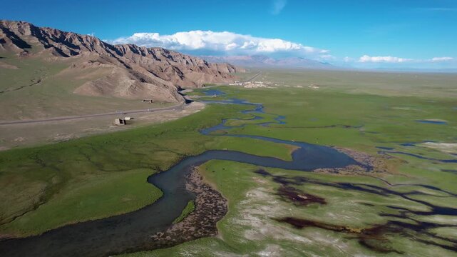 Aerial view of winding river through green grassland beside rocky mountains