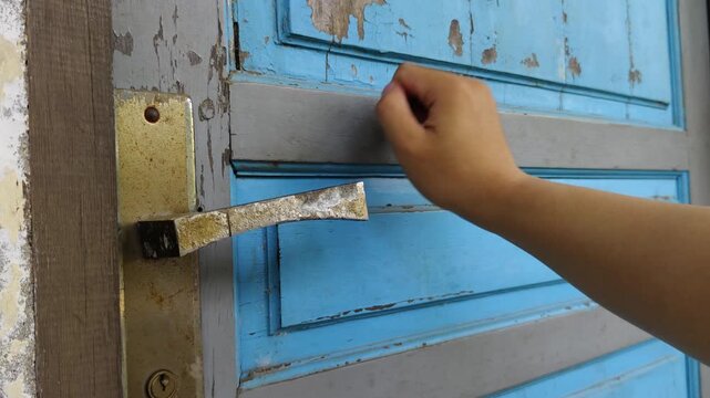 A close-up shot of a hand knocking on a weathered, dark blue wooden door&mdash;with peeling paint and rusty hinges&mdash;in natural light.