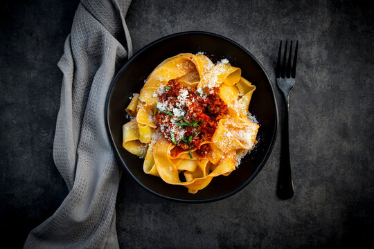 Bowl of pappardelle pasta with bolognese sauce rests on dark concrete