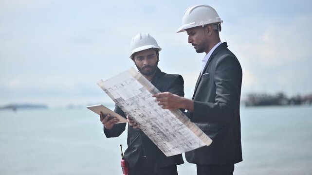 Engineers wearing safety gear, including hard hats examining survey a large blueprint tablet standing industrial facility gas or oil refinery engaged in a job requires high safety standards concept.	