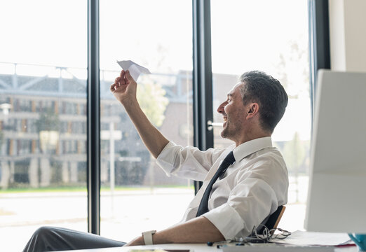 Smiling businessman sitting at desk in his office looking at paper plane