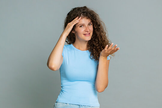 Young woman making facepalm gesture covering face with hand, laughing humorously at silly, funny mistake. Girl on gray background expresses humorous reaction to embarrassing misunderstanding situation