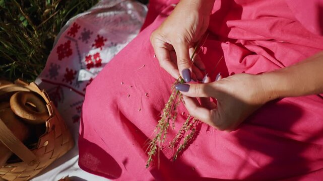 Close up of woman hands arranging wild grass on bright pink fabric with traditional embroidered cloth woven basket containing bread and bagels croissant visible. Soft natural light