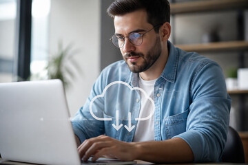 Fototapeta premium A young man wearing glasses and a denim shirt is working on a laptop in an office setting, with a cloud icon overlay suggesting data or connectivity.