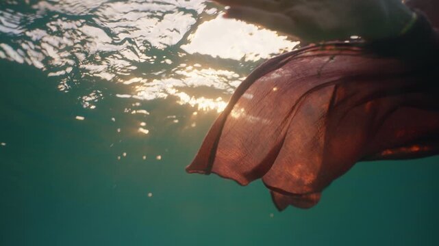 A woman swims toward the camera underwater, her dress flowing as sunlight beams down.