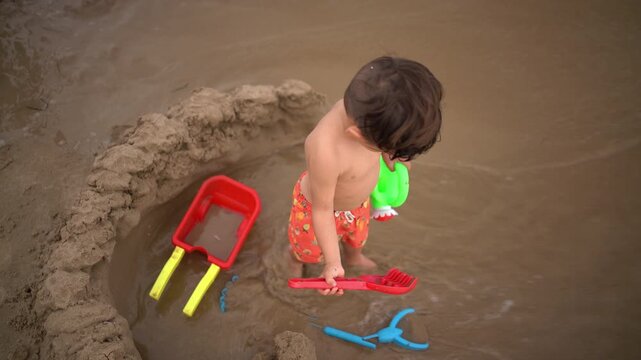 An overhead shot shows a toddler holding beach tools as a wave washes into the hole he dug, soaking him and his toys