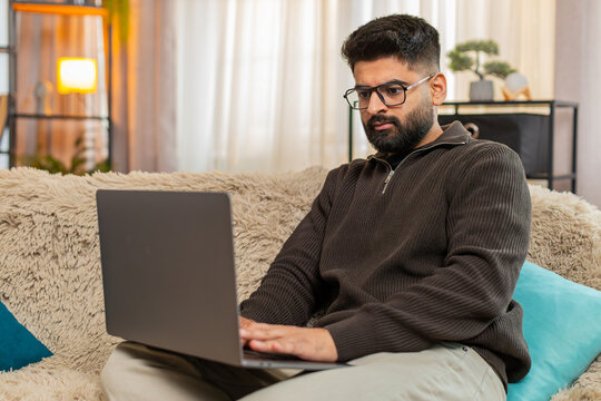 Indian man at home types on laptop with focused expression, preparing for upcoming online exam with full attention. Arabian guy on sofa concentrates on studying and memorizing materials for success