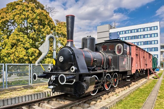Historic black steam locomotive engine with red wheels and wooden cargo wagon displayed outdoors on tracks under a blue sky. Concept of railway history, vintage transport, and industrial heritage.