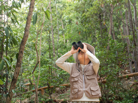 Little Asian girl wearing safari hat and vest looking through binoculars on forest nature trail
